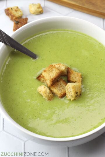 White bowl filled with green potato zucchini soup and croutons.