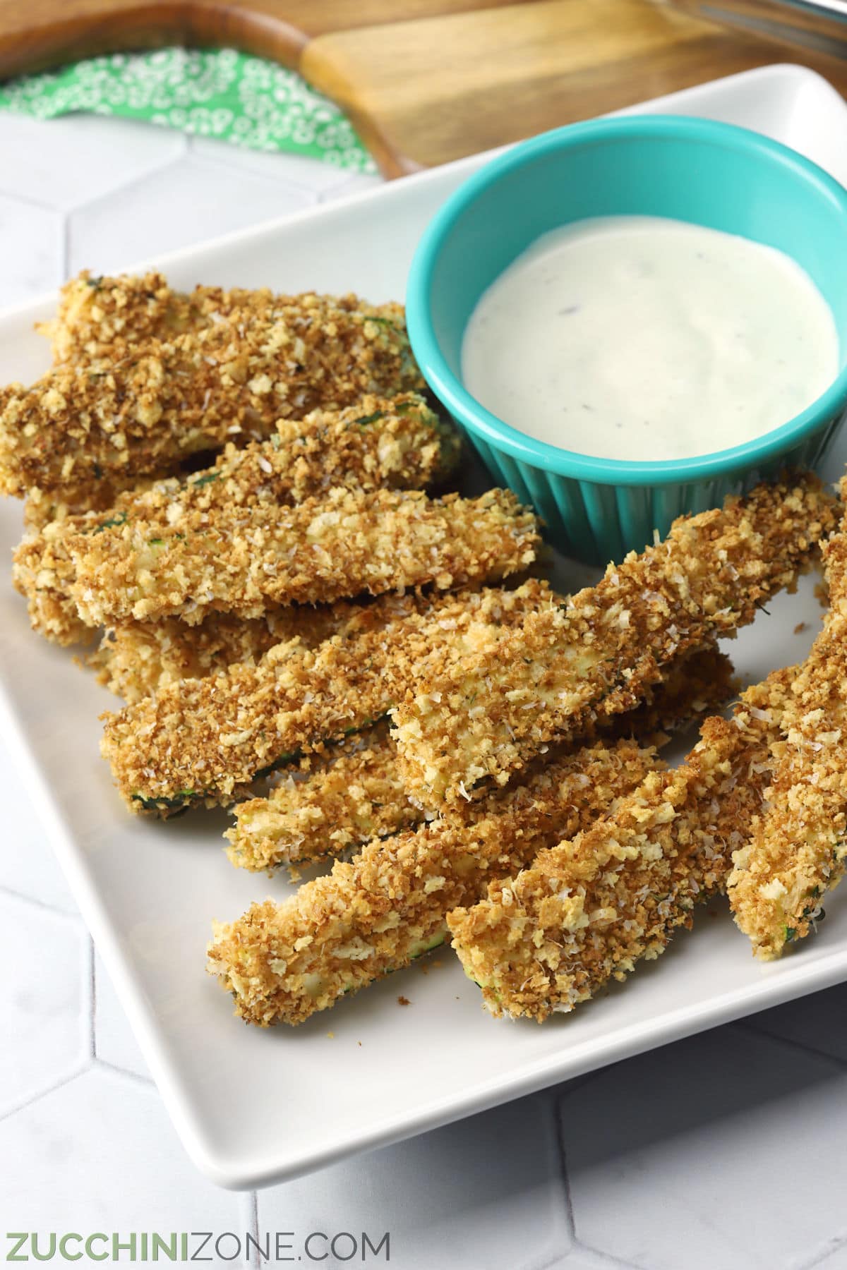 A square white plate filled with baked zucchini sticks and a teal bowl of ranch dressing.