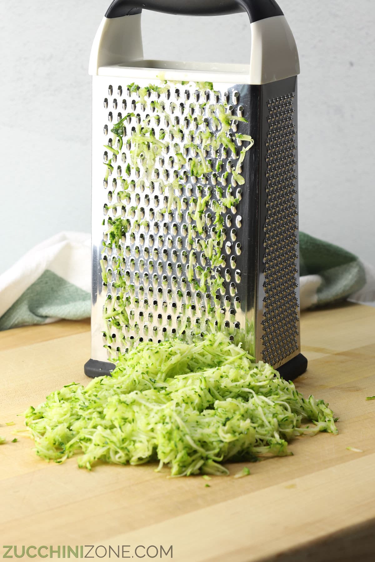 Shredded zucchini in front of a box grater on a wooden cutting board.