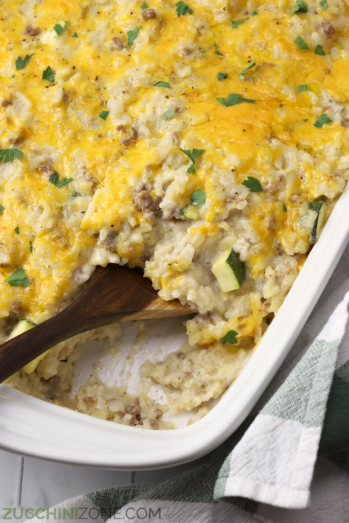 Wooden spoon scooping cheesy zucchini casserole from a white casserole dish.