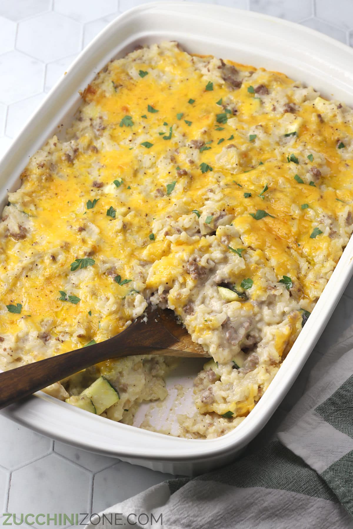 A white casserole dish filled with cheesy rice casserole with a wooden spoon.