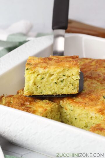 A slice of cornbread zucchini casserole sitting on a spatula on top of the remaining casserole.