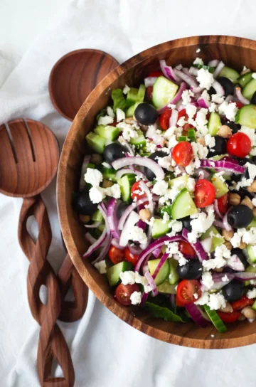 A wooden bowl with wooden serving utensils filled with Greek salad.