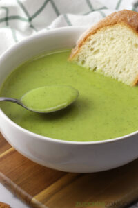 Close up of bowl of green soup with a metal spoon and a slice of bread.