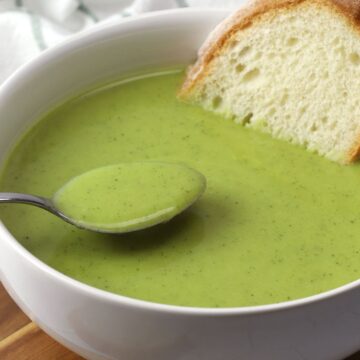 Close up of bowl of green soup with a metal spoon and a slice of bread.
