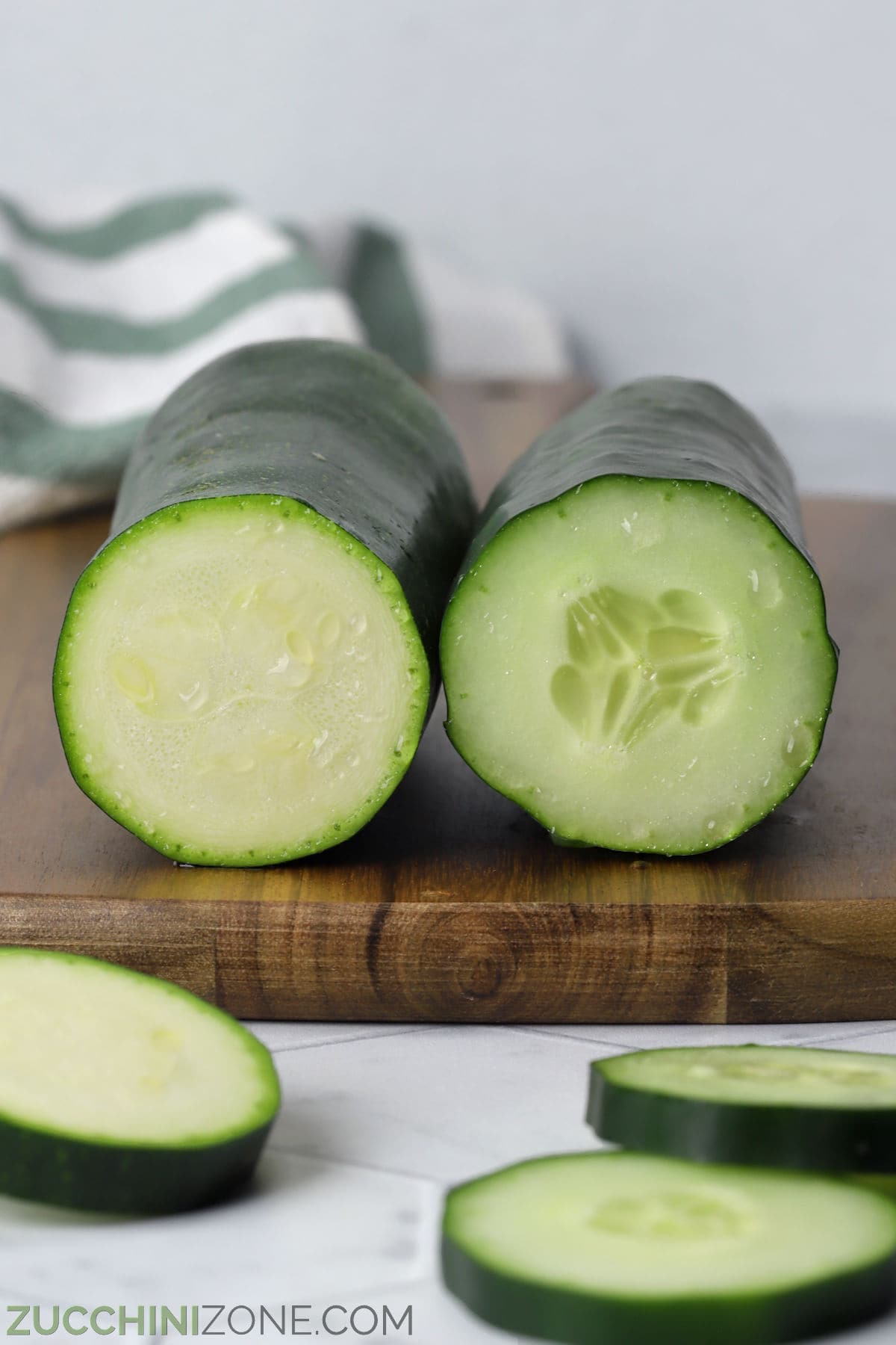 A sliced zucchini and sliced cucumber laying side by side on a wooden cutting board.