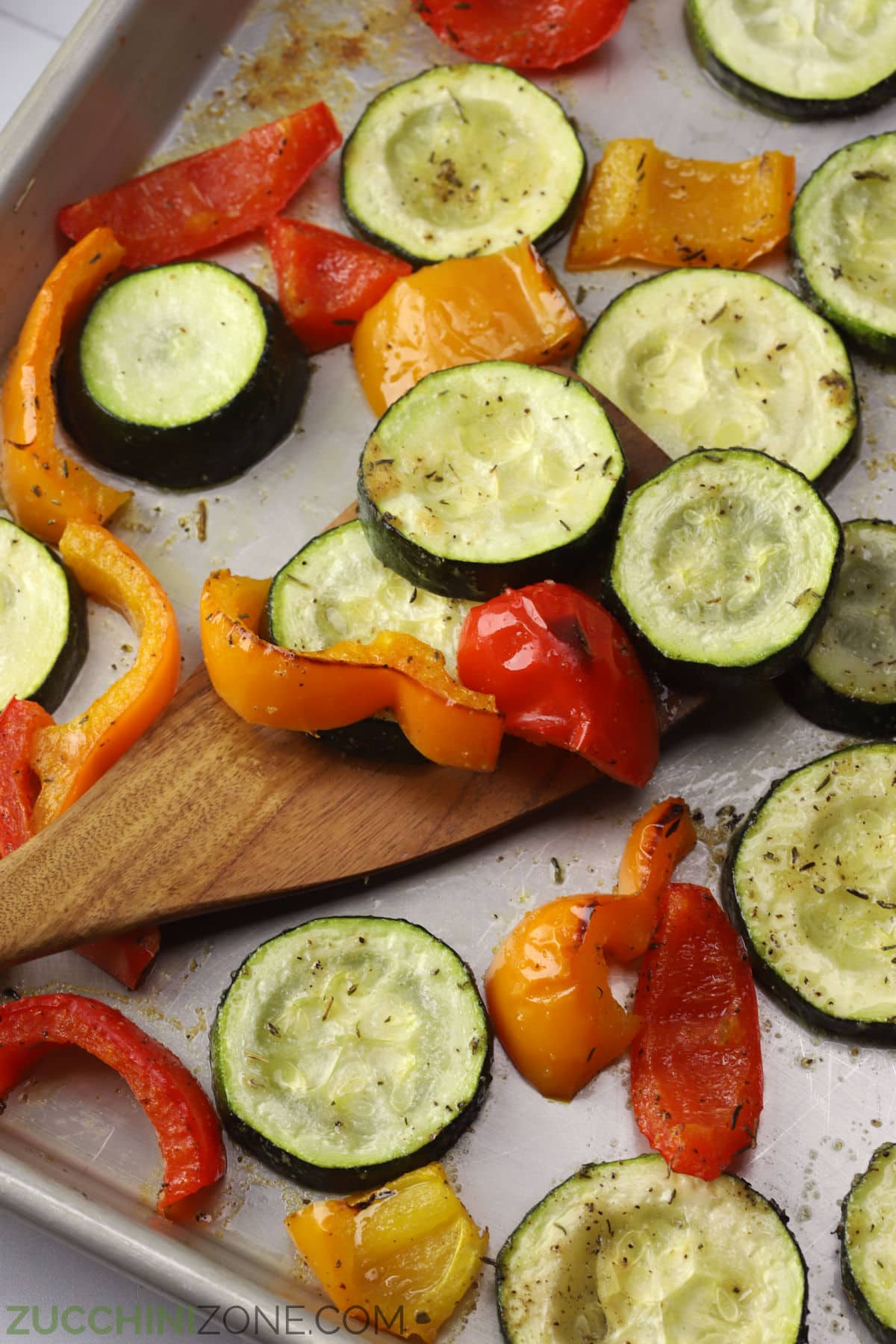 Wooden spatula scooping zucchini and peppers from a sheet pan.