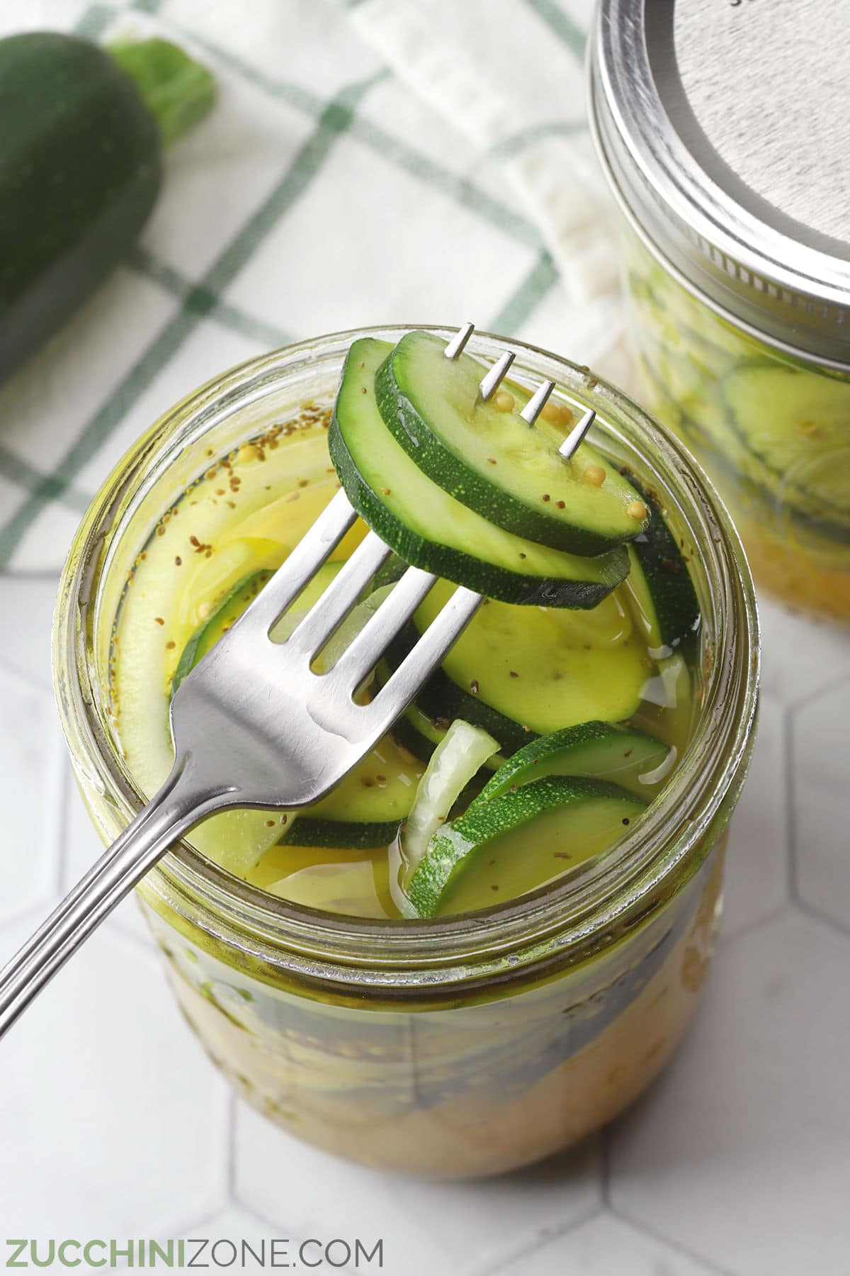 A fork piercing several zucchini pickles in an open pickle jar.