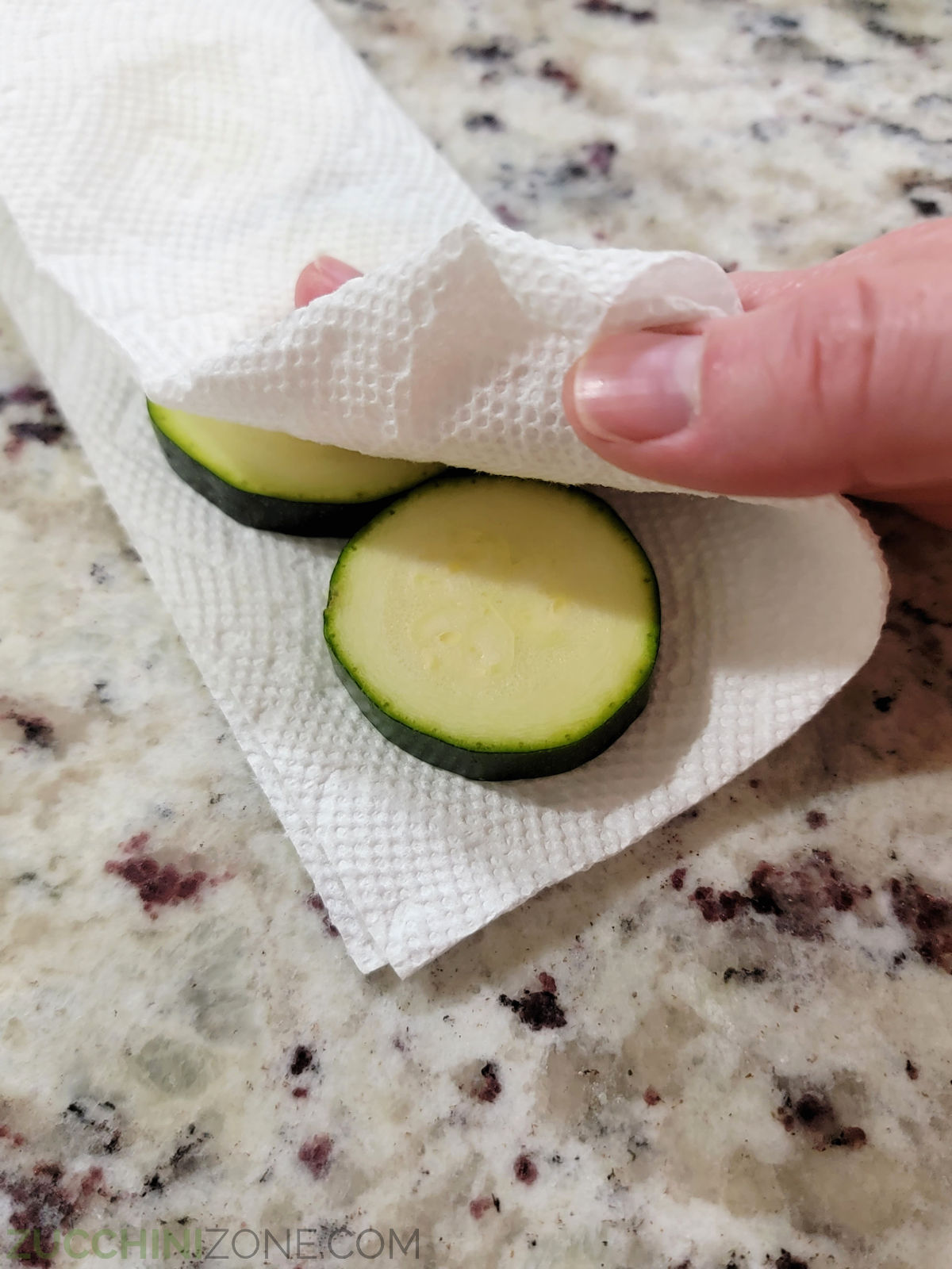 A hand patting zucchini slices dry with a paper towel.