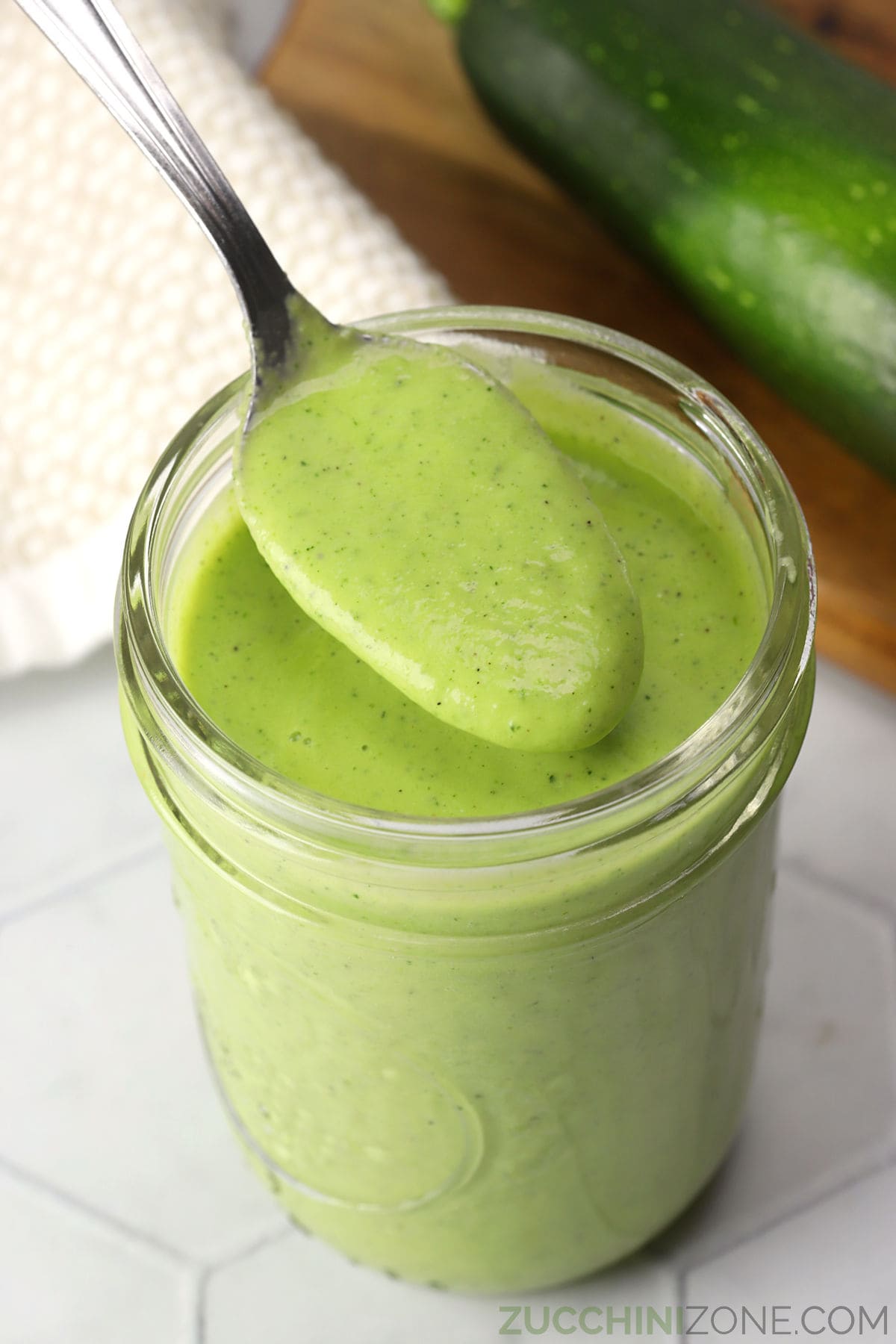 Close up of a spoon held over a glass jar filled with green zucchini salad dressing.